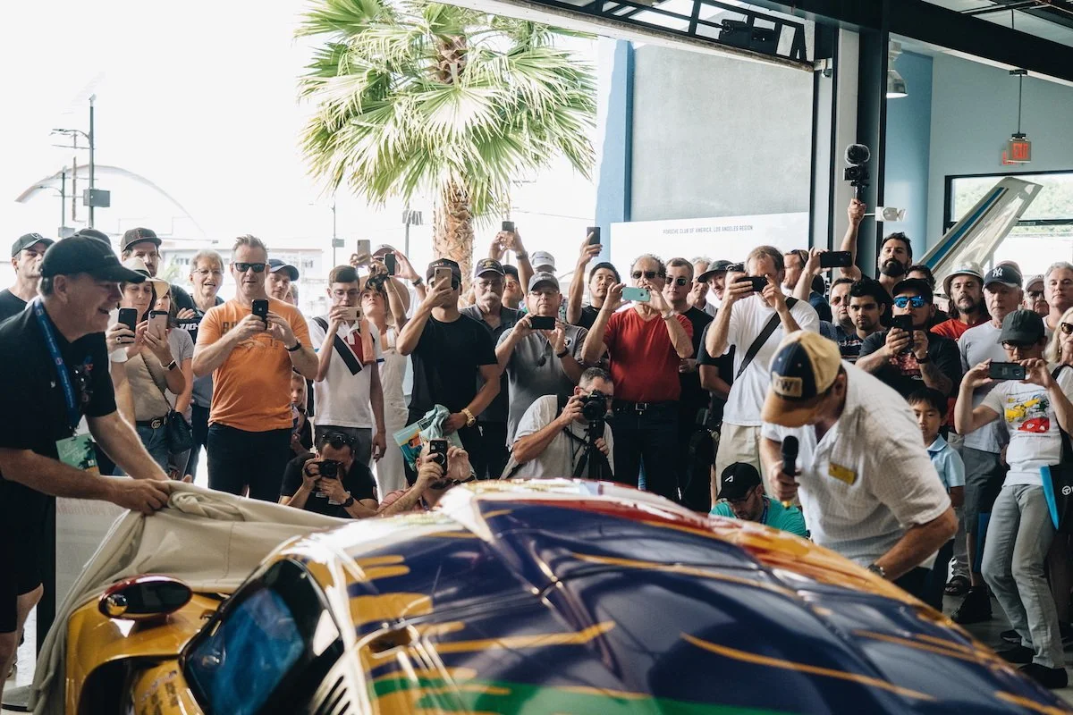 A crowd of people taking photos and videos at an event where a yellow sports car is being displayed indoors. The scene includes a palm tree and a man with a microphone and a white shirt.
