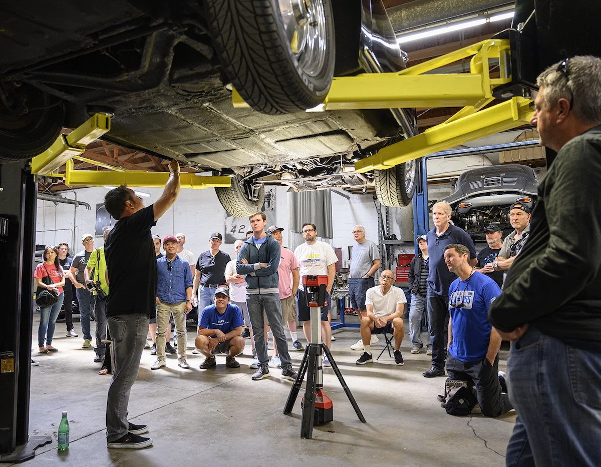 A man is demonstrating under a car on a hydraulic lift to a group of people in an automotive workshop. The audience is watching attentively.