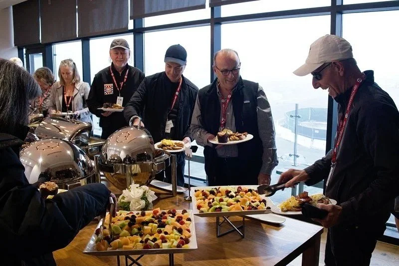 People serving themselves buffet-style food at a table with fruit and breakfast dishes, indoors with large windows.