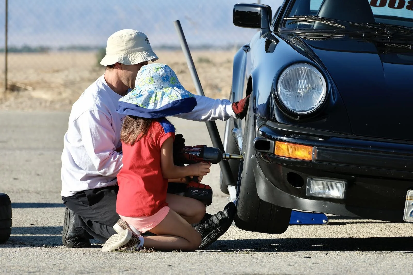 An adult and a young girl working on a black sports car with a jack under the front wheel in a desert area.