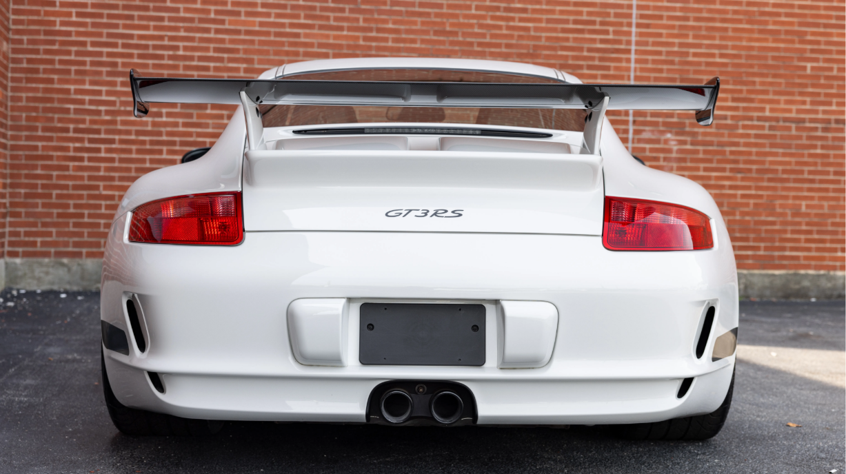 Back view of a white Porsche GT3 RS sports car with a large rear spoiler, parked in front of a red brick wall.
