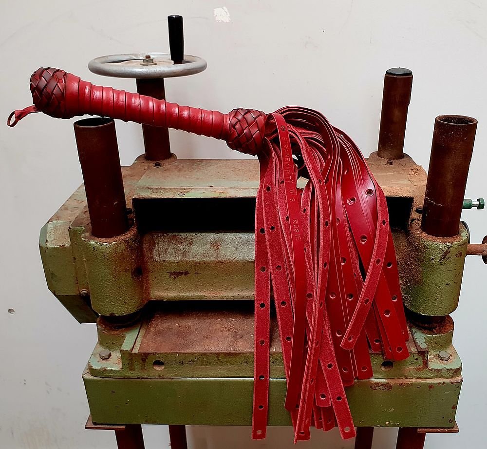 A vintage leather strap-making machine with multiple red leather strips draped over it, featuring a woven leather handle, on a white wall background.