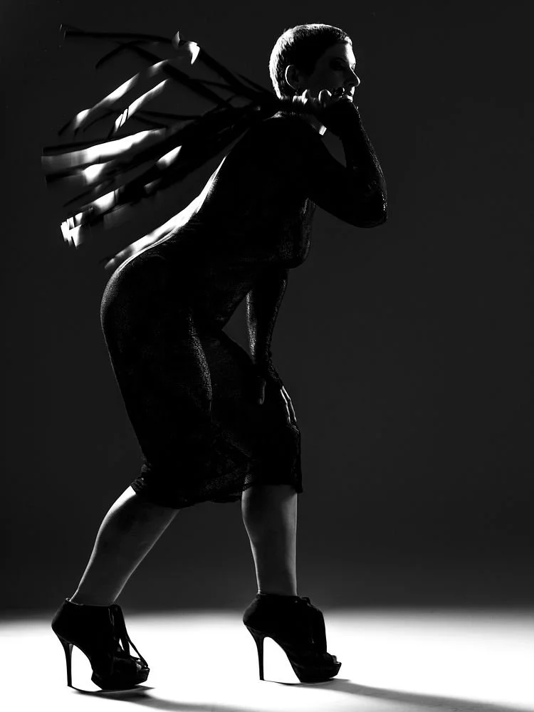 Silhouette of a woman with high-heeled shoes, wearing a dress and holding sunglasses, against a dark background.