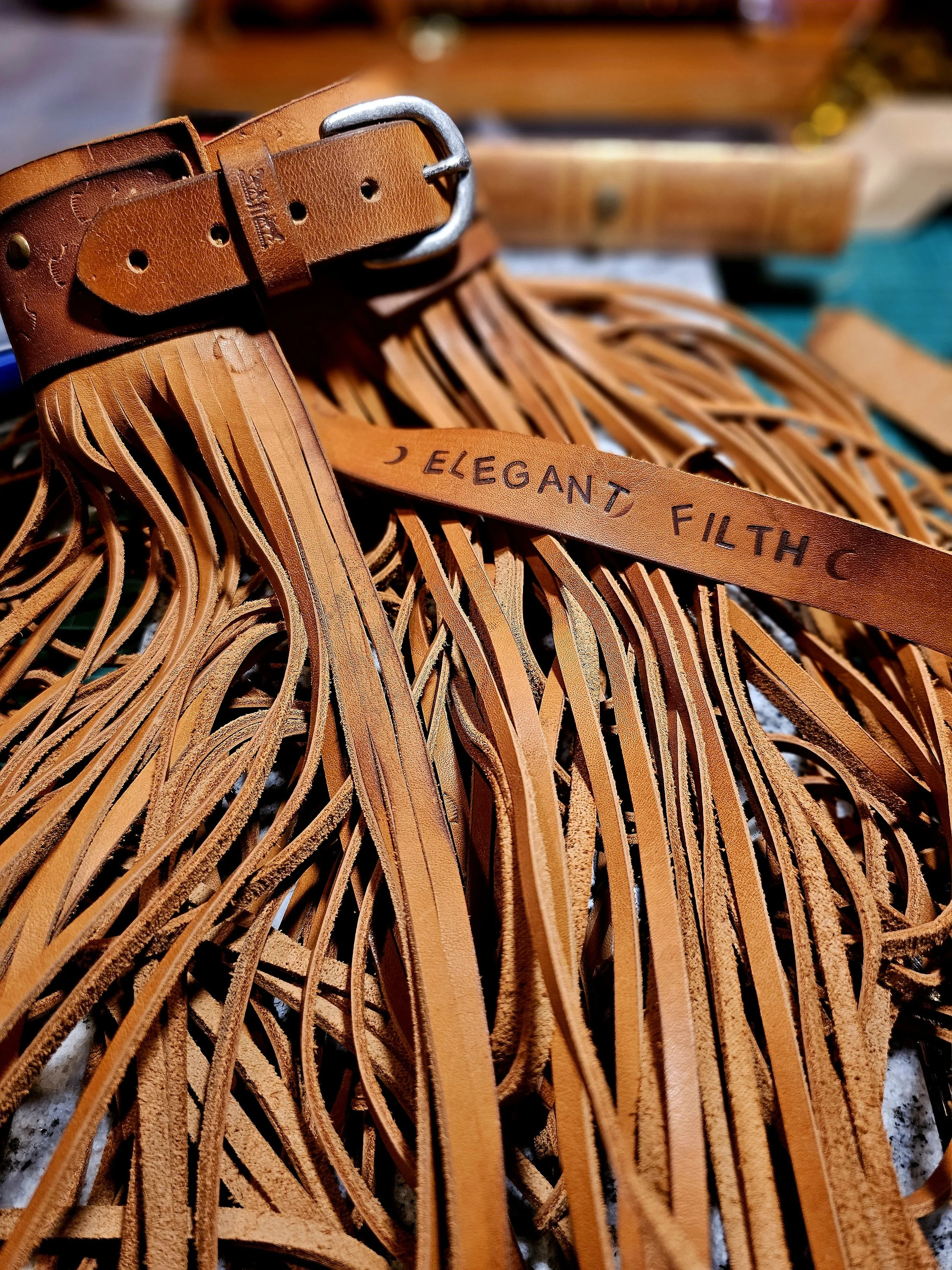 Close-up of a brown leather fringed accessory with a buckle, and a strap inscribed 'ELEGANT FILTH'.