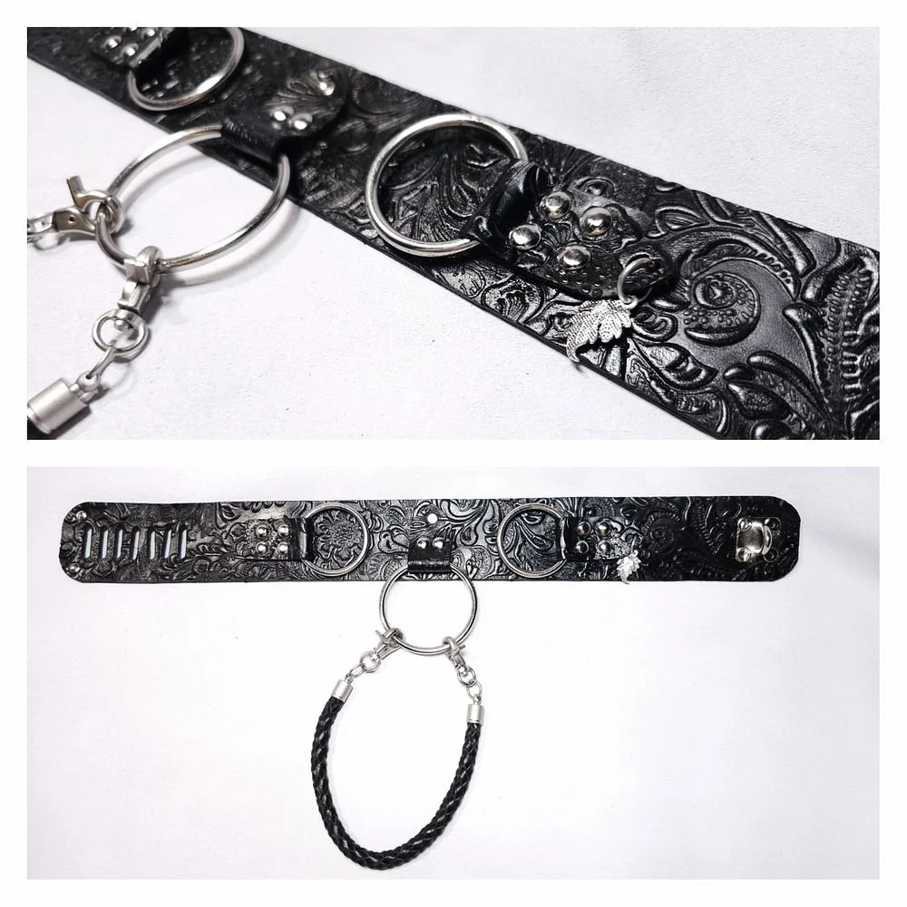 Close-up of a black embossed leather belt with metallic rings, studs, and a detachable chain link. The belt is laid flat on a white surface, showing intricate floral and paisley patterns.