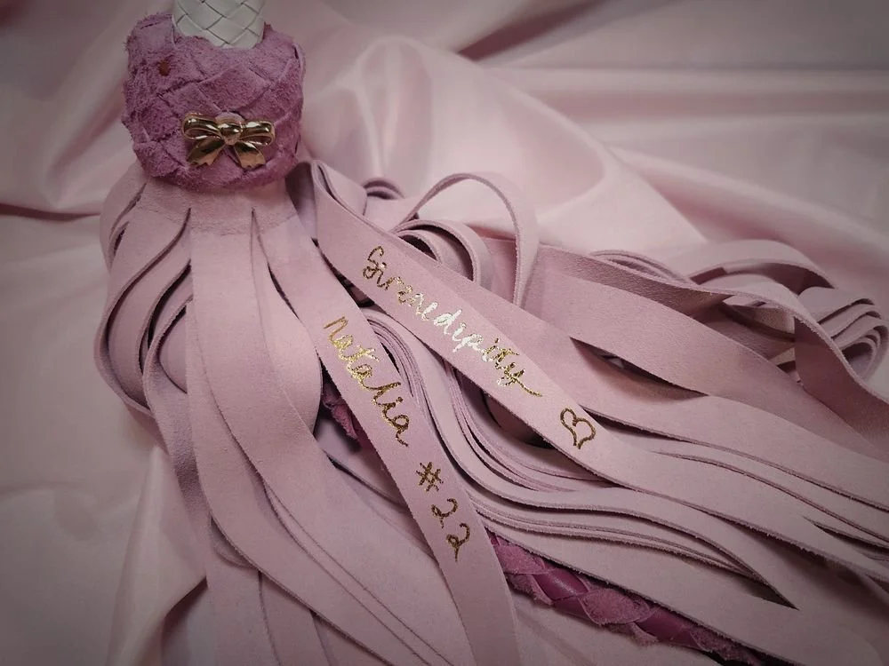 Pink decorative item resembling a layered ribbon with a small pink sock, adorned with a gold bow, and handwritten text on ribbons, placed on pink fabric.