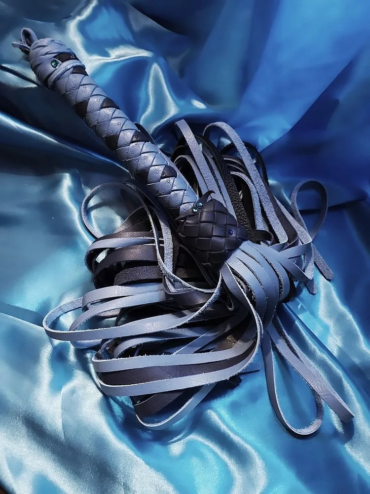 A leather flogger with a braided handle resting on a shiny, satin blue fabric.