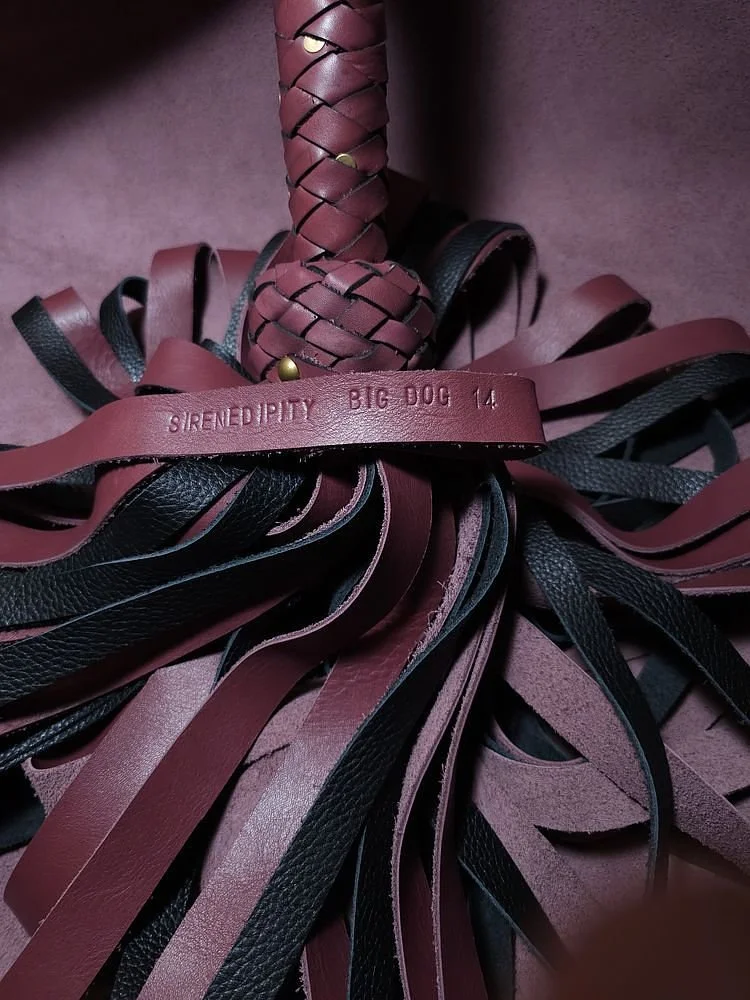 Close-up of a bundle of leather strips in black and maroon, with a leather strap embossed with 'SIRENDIPITY BIG DOG 14' and a braided leather handle.