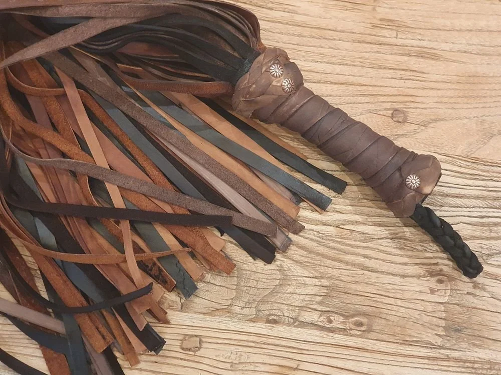 A leather-wrapped handle with metal decorative studs, attached to a woven leather tassel with strips of leather in brown, tan, and black colors, lying on a wooden surface.