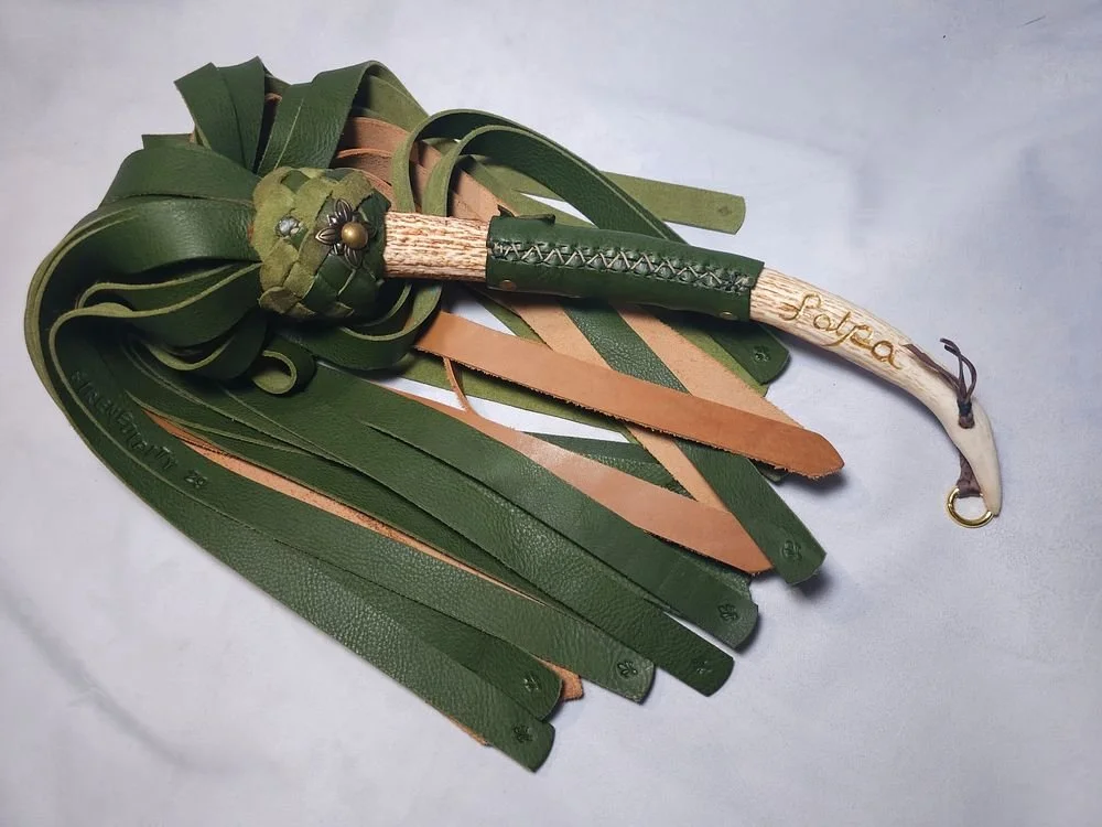 A leather whip with multiple green and tan straps, having a decorated handle with an engraved name, lying on a light surface.