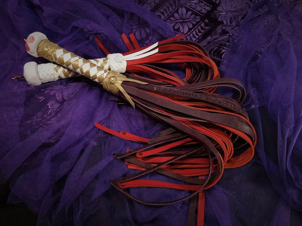 A handmade whip with a handle decorated with gold and silver accents and multiple leather strips in red and dark purple, lying on purple fabric.