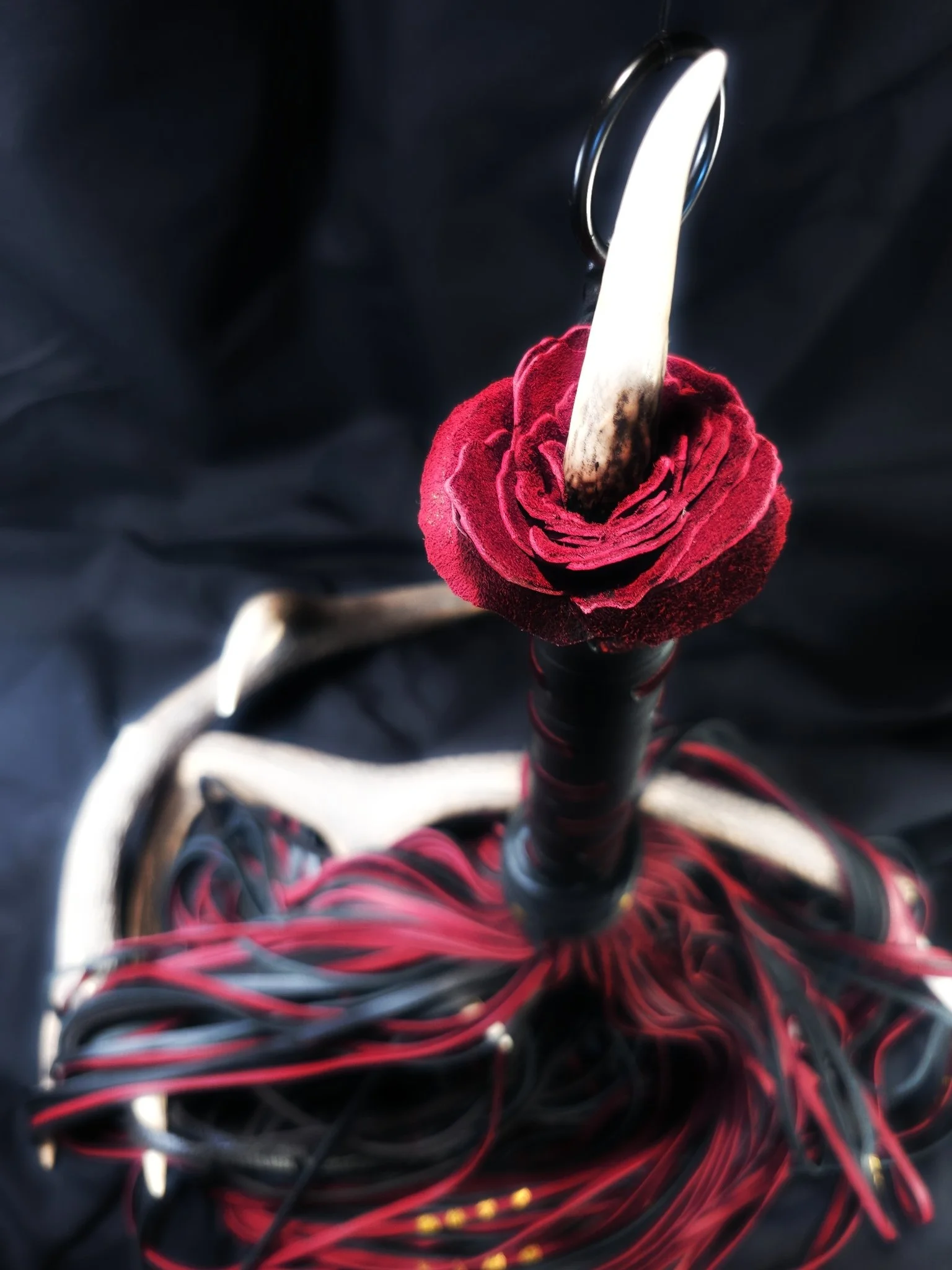 A close-up view of a fantasy sword with a red velvet rose at the hilt, surrounded by black and red braided leather cords.