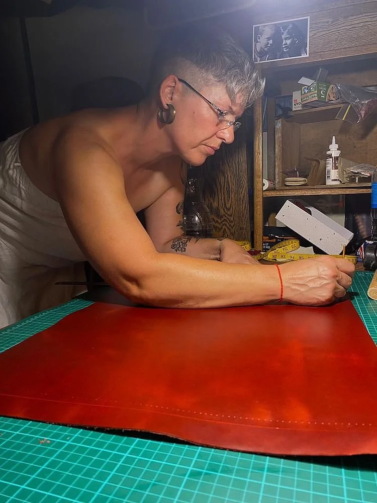 A woman with short gray hair, glasses, large earrings, and a tattoo on her arm is working on a leather project at a green cutting mat in a workshop.