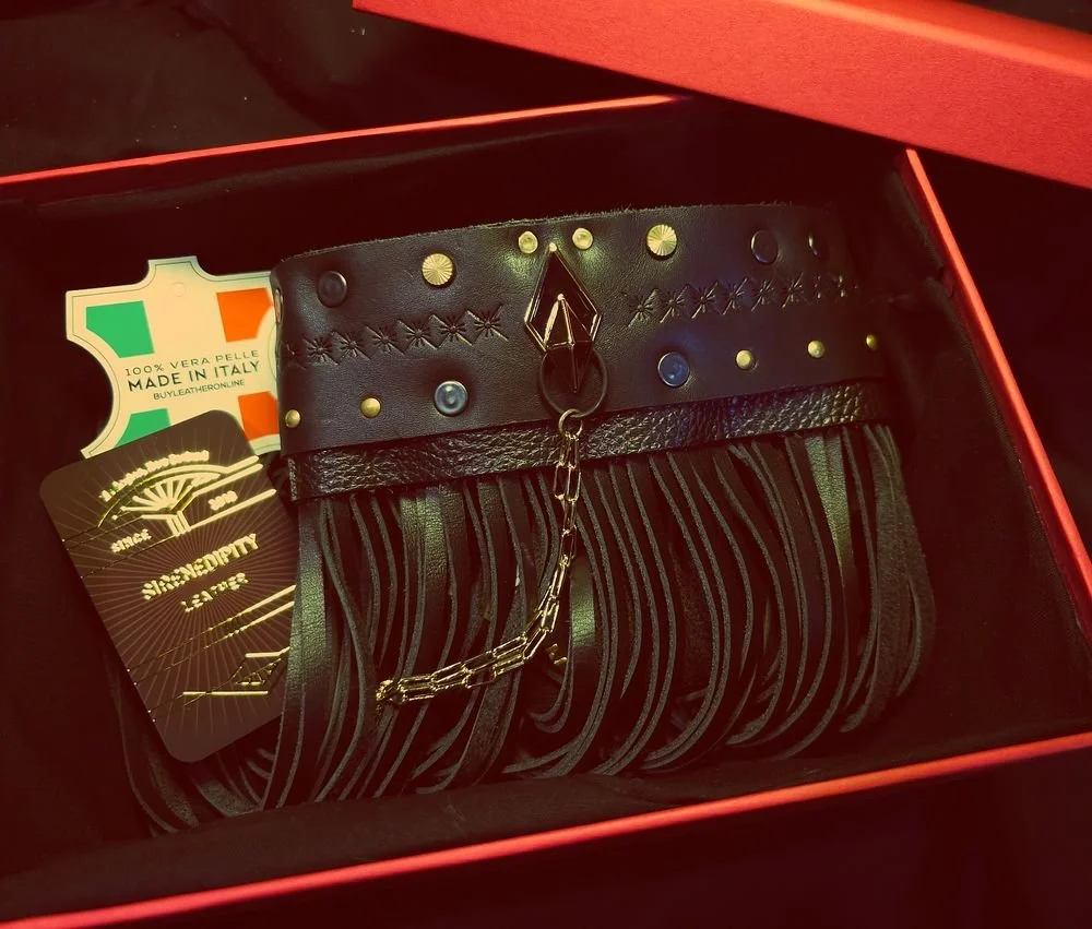 A black designer clutch purse with gold and metallic studs and a chain, inside a red box, with tags indicating it is Italian-made leather accessories.