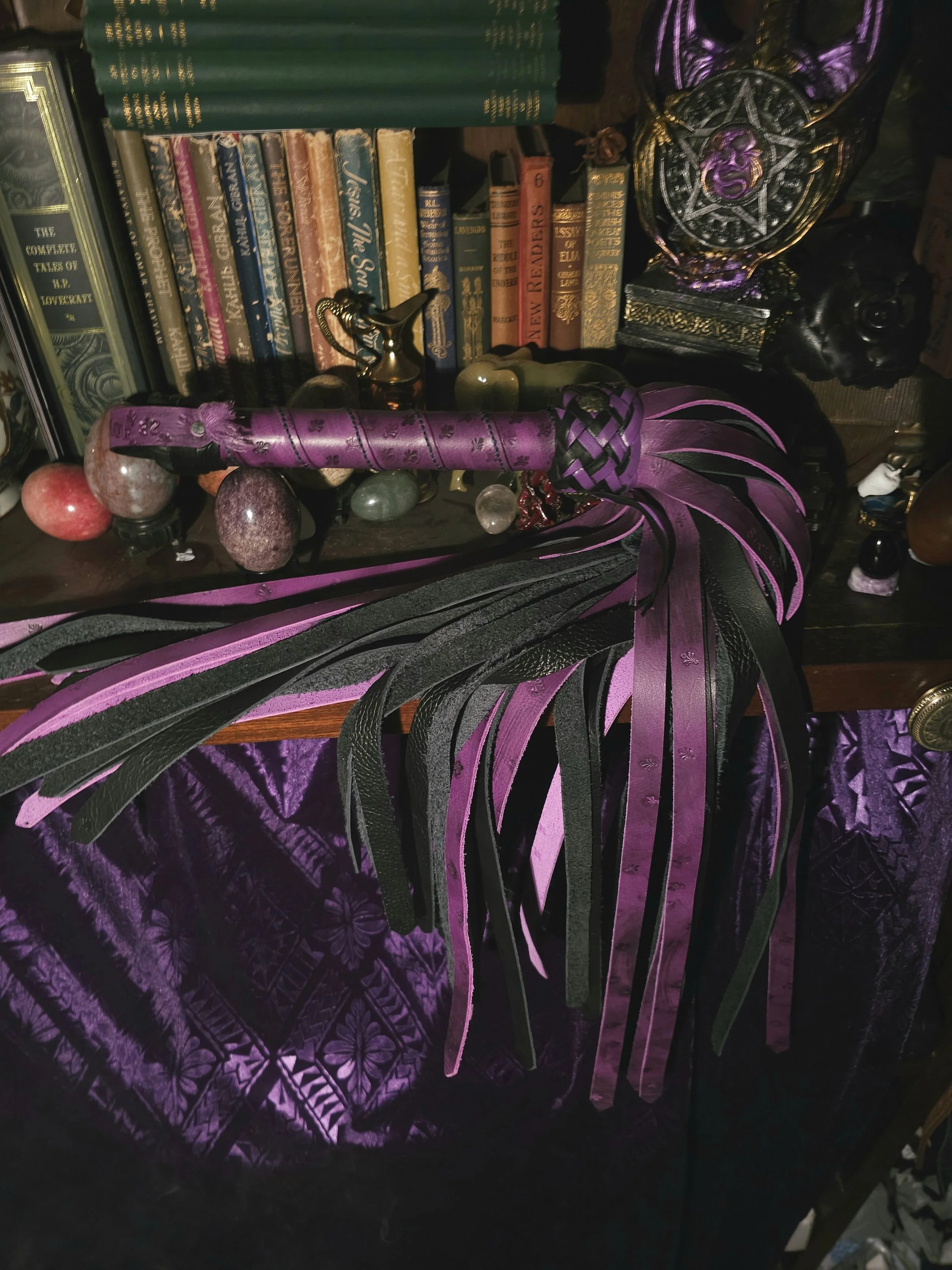 A purple and black leather flogger with long strips, a purple handle with a bow design, placed on a dark wooden surface in a room with books, crystals, and decorative objects.