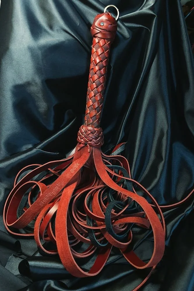 A leather flogger with a braided handle and multiple leather tails resting on black fabric