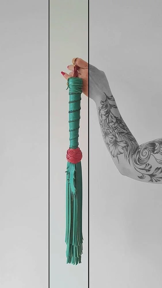 A tattooed arm holding a turquoise flogger with a pink handle against a plain white background.
