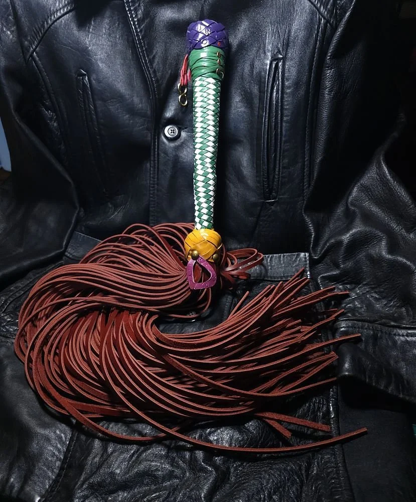 A whip with a colorful handle and long brown leather strands, resting on a black leather jacket.