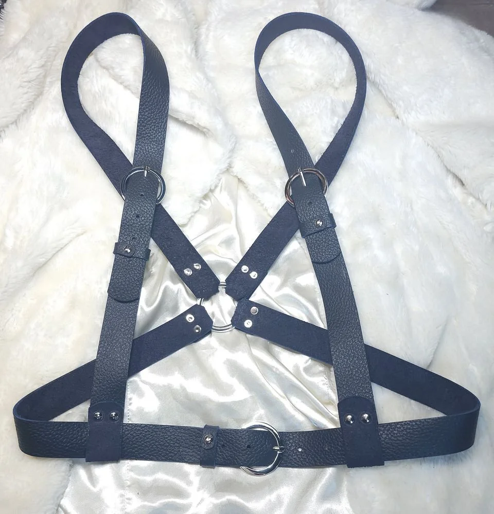 Black leather harness with silver metal rings laying on a white plush surface.