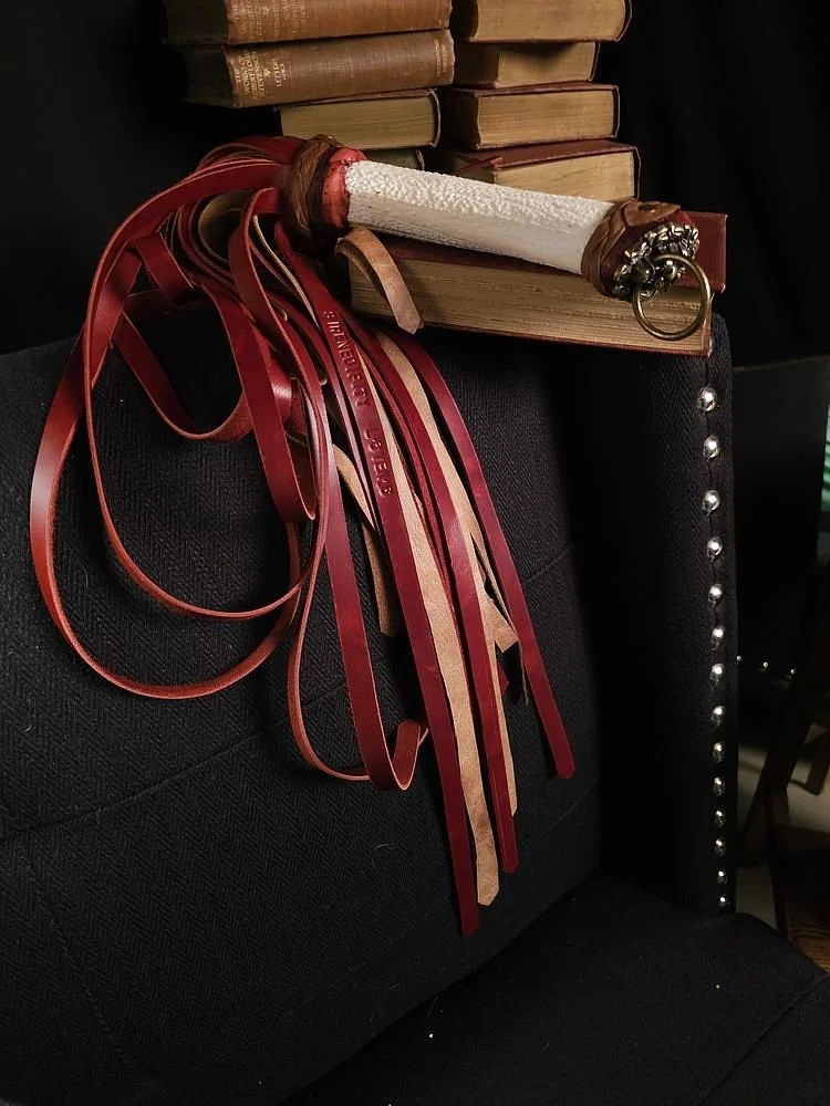 A black chair with decorative silver studs, stacked old books on top, and a sword with a white handle and red ribbons hanging from the hilt resting on the books.