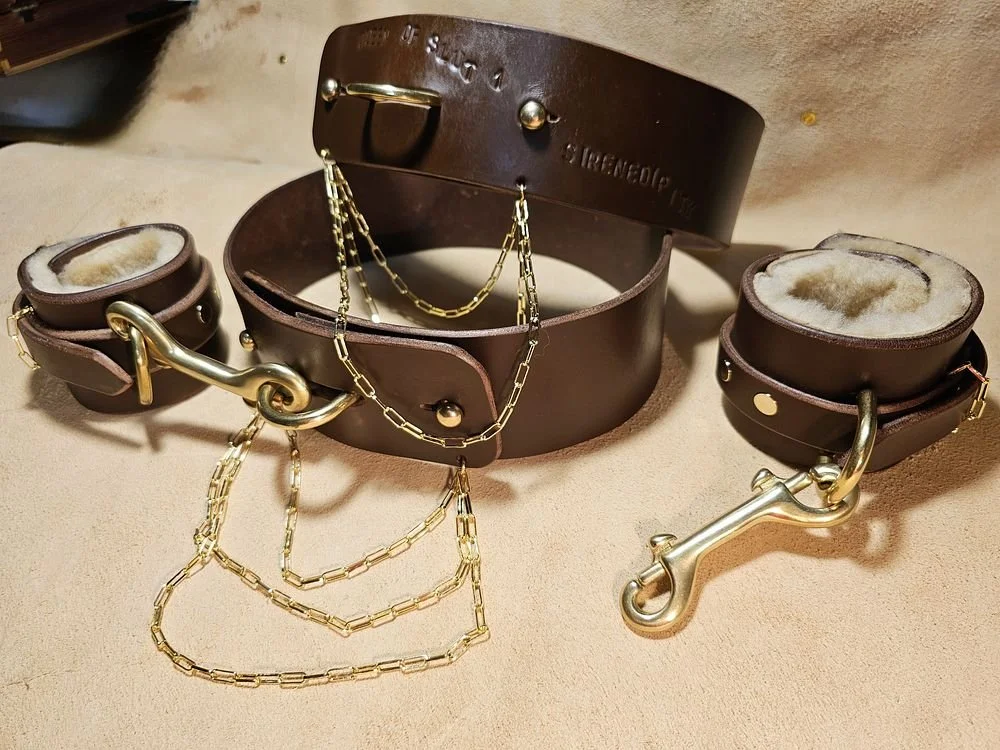 A leather dog collar with matching cuffs and chains, with a large hook on each cuff, on a beige surface.