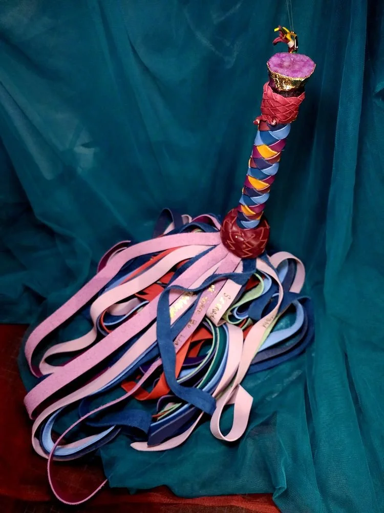 A colorful artistic sculpture resembling a gavel, with a handle wrapped in multicolored ribbon-like material and a pink top, surrounded by a pile of variously colored ribbon strips on a blue fabric background.