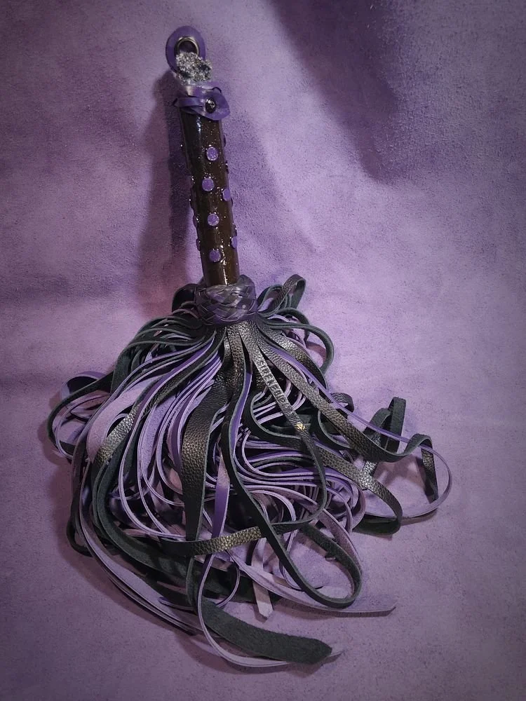 A whip with a purple handle and multiple black and purple leather straps hanging from the handle, against a purple background.