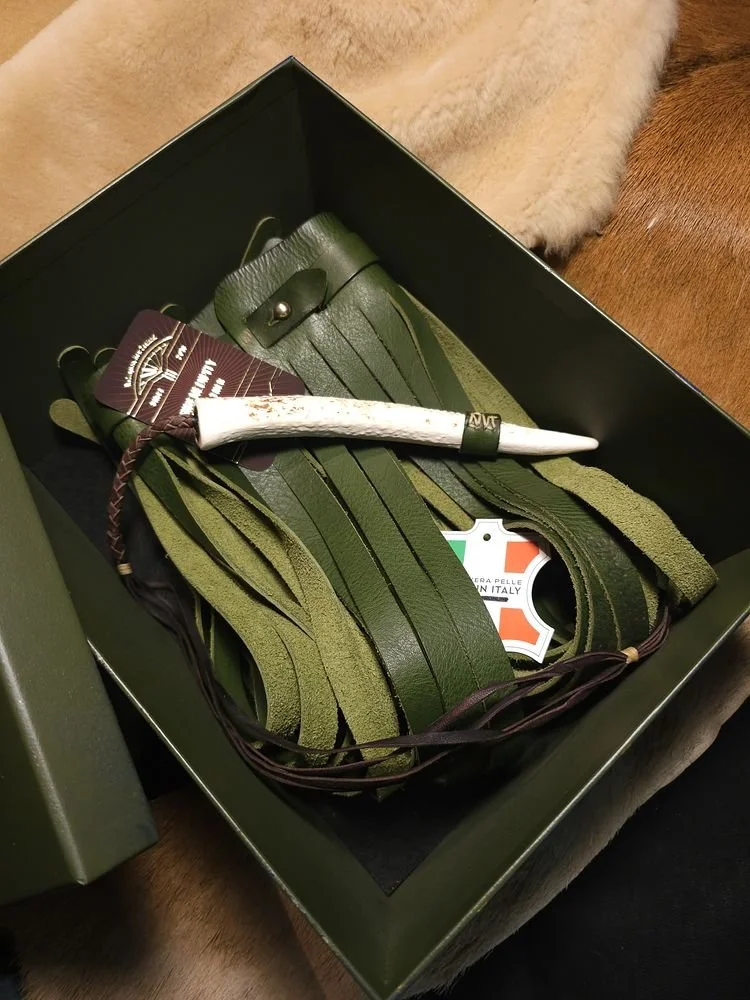 A box containing a leather watch strap, a white knife with a textured handle, and some small tags or packaging labels, resting on a blanket and wooden surface.