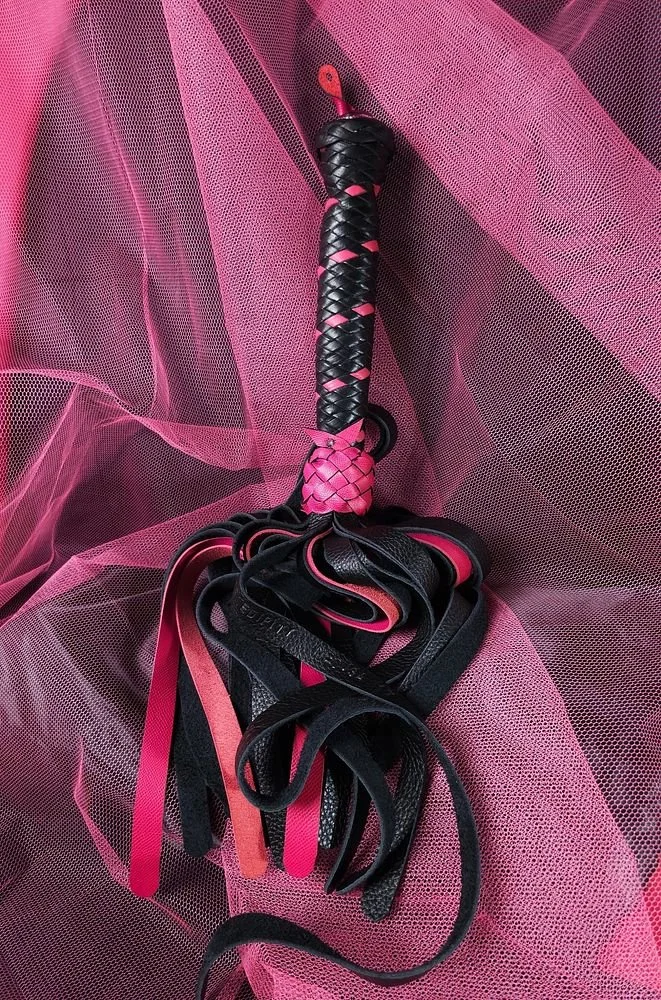 Whip with a braided handle, wrapped in black and pink, resting on pink mesh fabric.