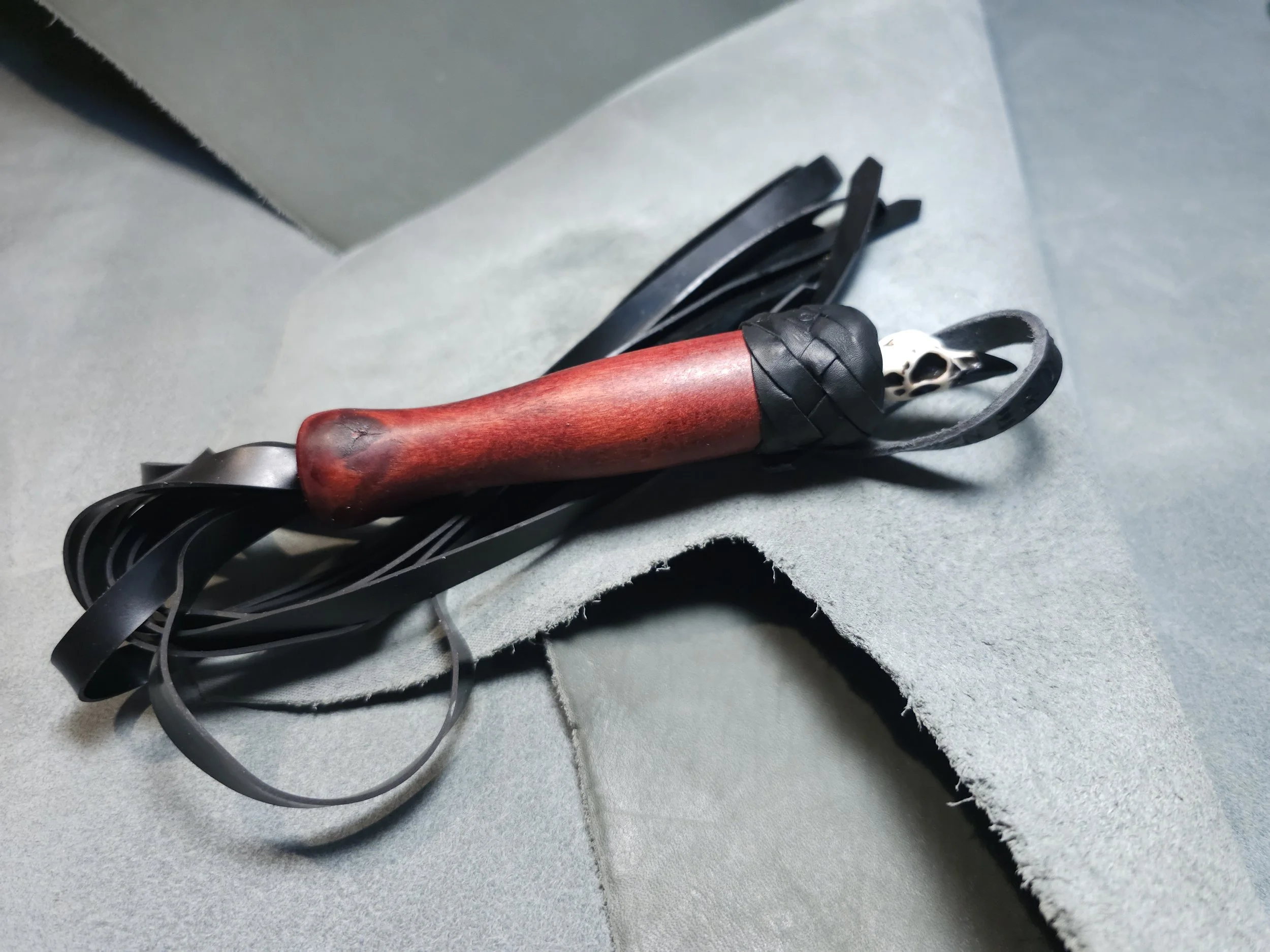 A leather riding crop with a wooden handle, a small animal skull, and a band of black leather, resting on a light gray surface.