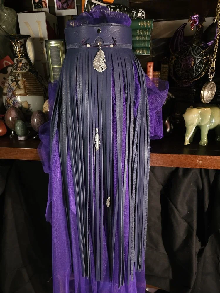 A purple and black leather fringed skirt with metallic leaf-shaped jewelry on a display stand surrounded by decorative objects, books, and collectibles.