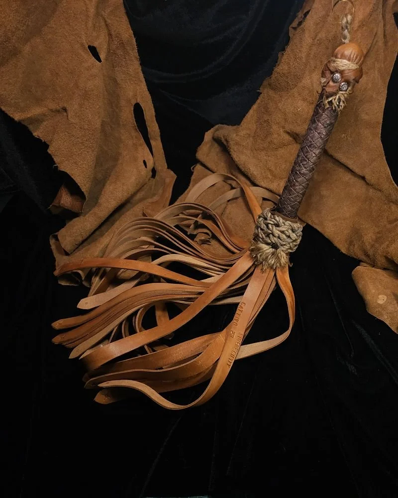 A leather whip with a decorative handle and a braided rope at the base of the handle, resting on a tan coat and black pants.