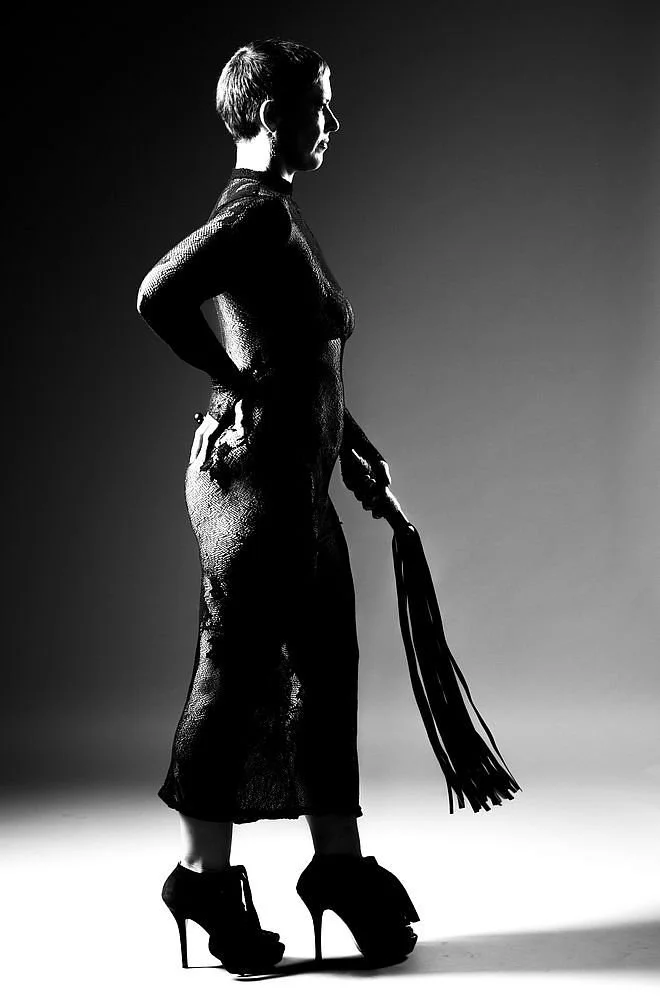A black and white photo of a woman with short hair, wearing high heels and a long dress, holding a whip, standing in profile against a plain background.
