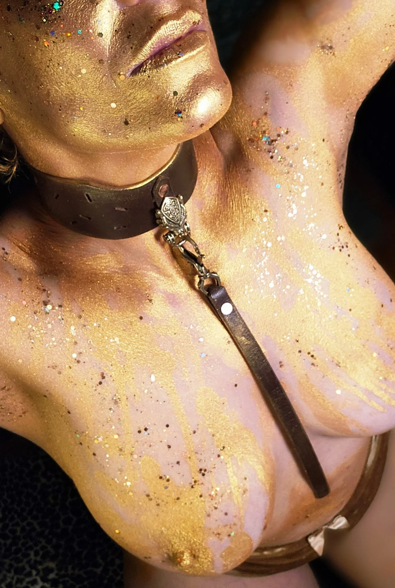 Close-up of a person with golden and glittery body paint, wearing a black choker with a metallic chain detail.