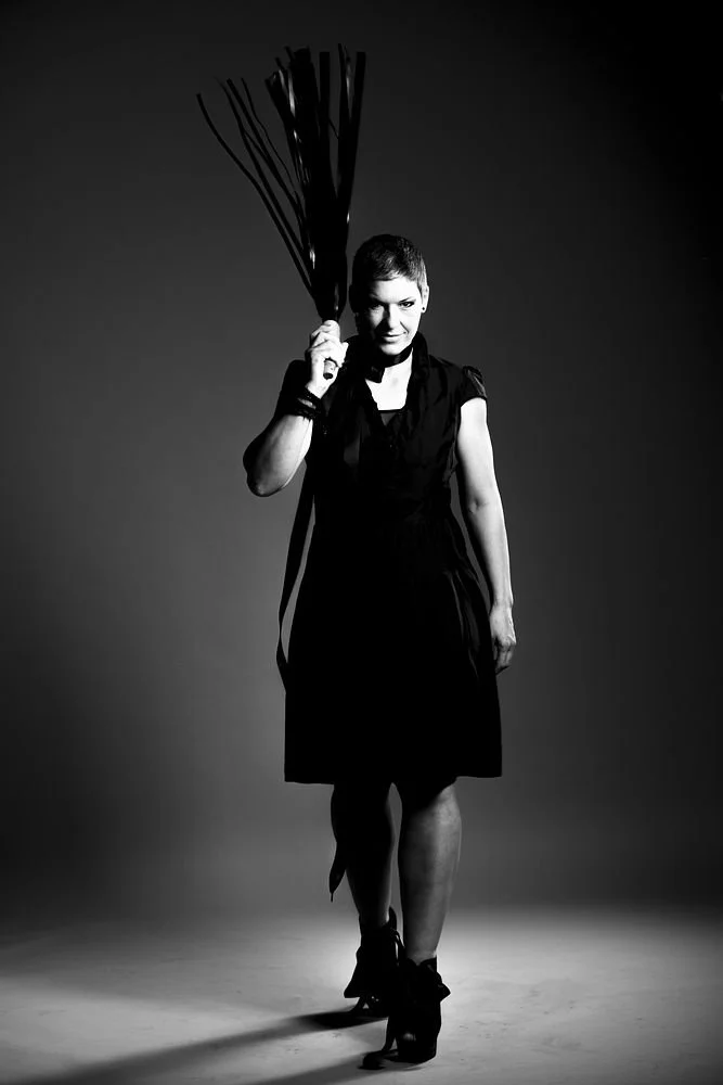 A woman dressed in black holding a bundle of sticks or twigs on her shoulder, standing in a studio against a plain background, in black and white.