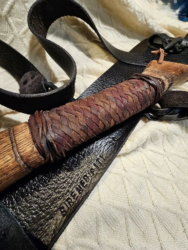 A close-up of a leather whip with a braided leather handle, a black leather thong, and a leather strap with the brand name 'SierreT' embossed on it, placed on a textured fabric surface.