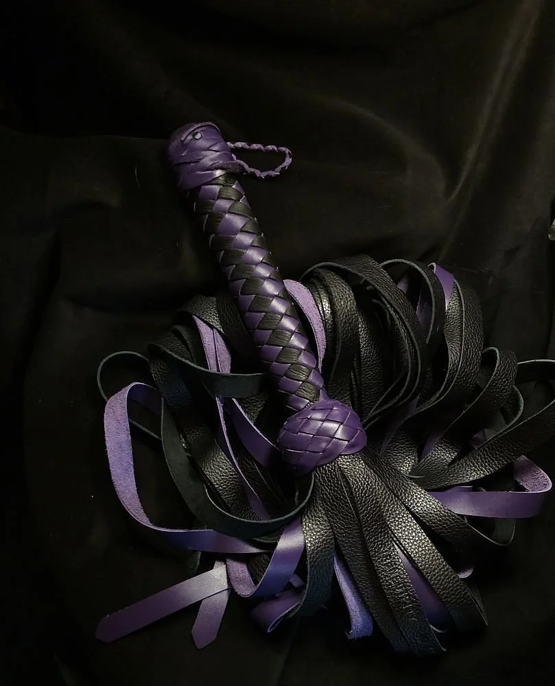 Purple leather whip with a braided handle resting on a pile of black and purple leather strips.
