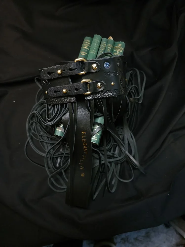 A black leather belt with gold studs and buckles, holding multiple green books and eyelash curlers, with black laces or cords hanging from it.
