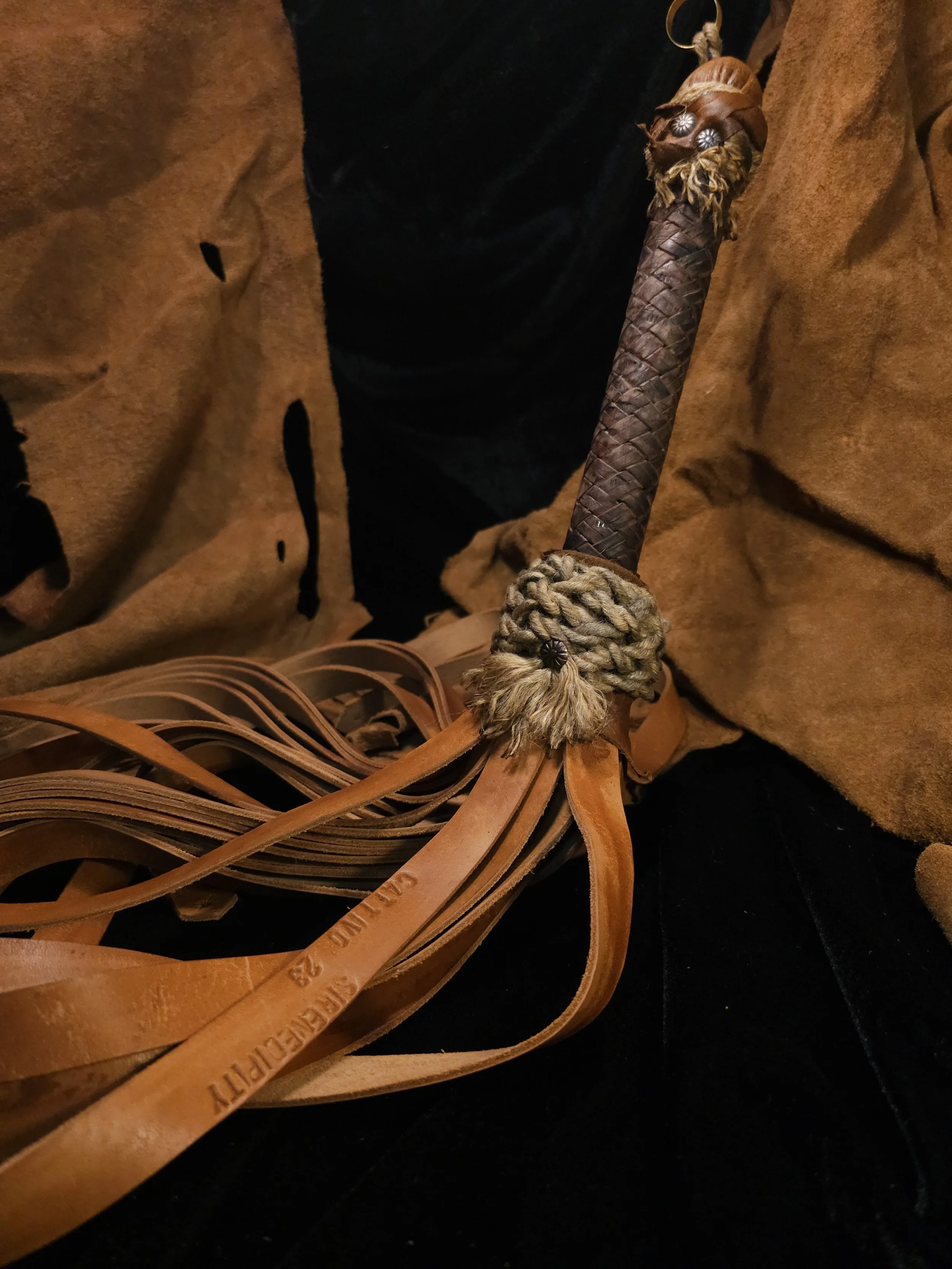 Close-up of a wooden or leather staff with woven leather wrap and decorative animal hair or fur, attached with metal studs or beads, held by a person in a brown suede or leather garment.