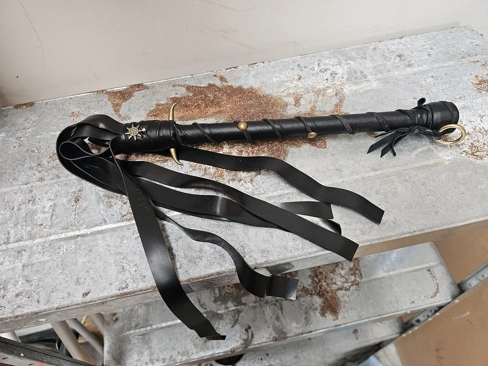 A decorative black leather whip with gold accents and black ribbons, resting on a weathered gray surface.