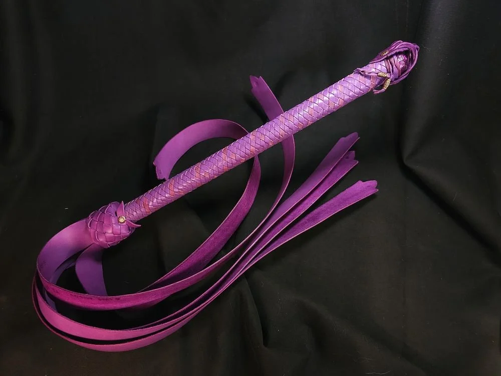 A purple whip with a braided handle and multiple long, thin strips, resting on a black surface.