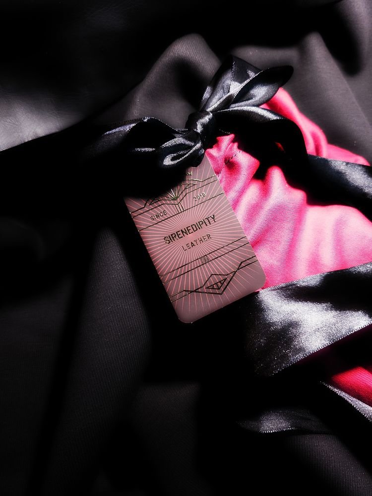 Close-up of a clothing tag that reads 'Sirenedepity Leather' attached to a garment with black satin ribbon and pink and black fabric.