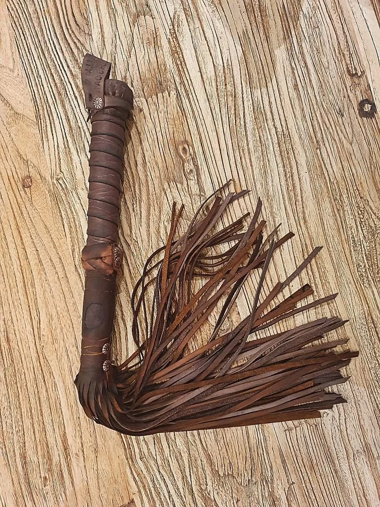 A handmade decorative item resembling a medieval-style hammer with a leather strap handle and a fringe made of leather strips, lying on a wooden surface.