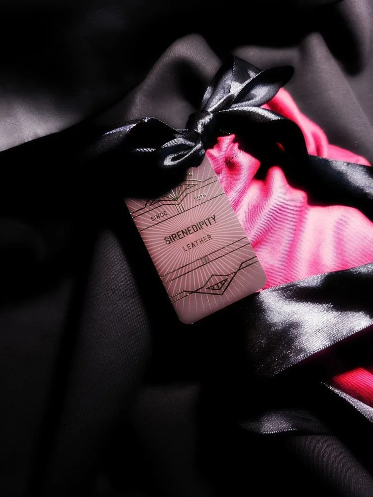 Close-up of a pink and black silk fabric with a tag that reads 'SURENEEDIETY LEATHER' attached with a black satin ribbon.