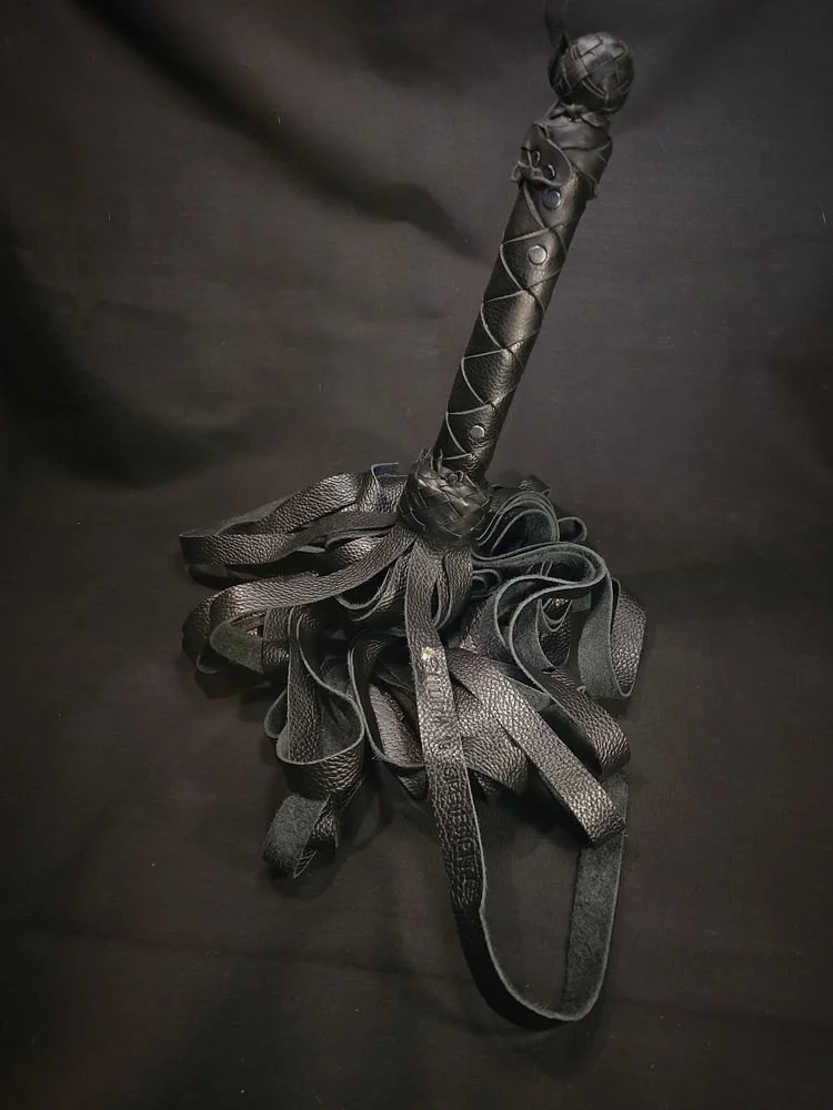 A black BDSM whip with multiple leather straps and a handle wrapped in black leather with metal studs, resting on a black surface.