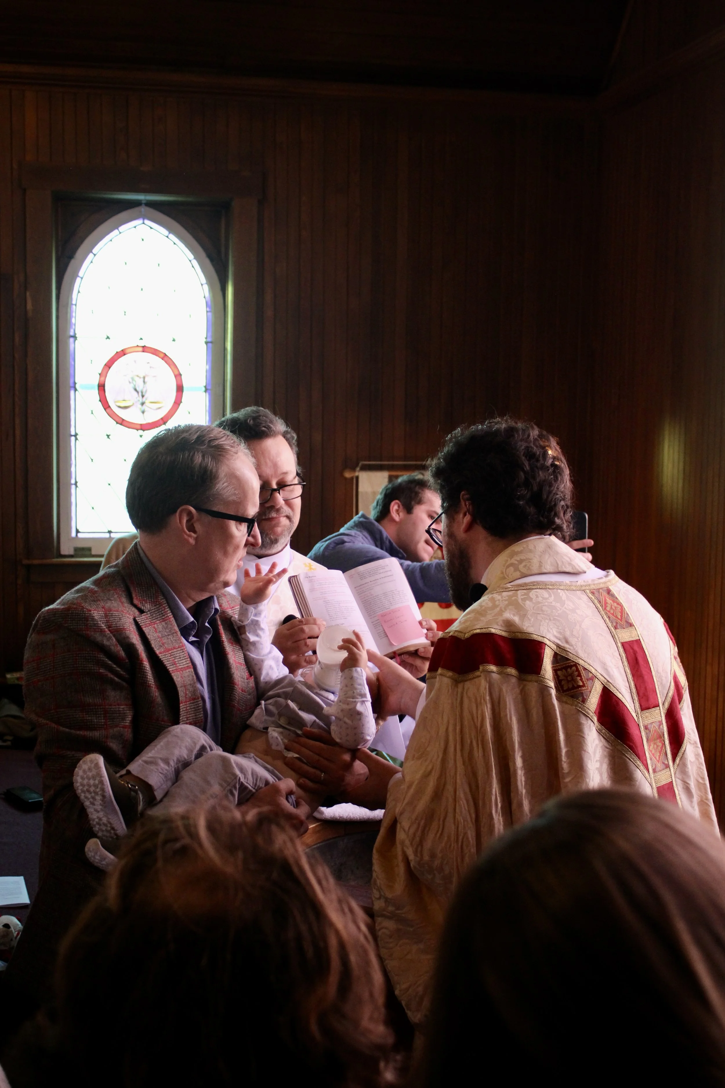 A religious baptism ceremony inside a wooden church with stained glass window, where a man is holding a baby and a priest appears to be performing the baptism.