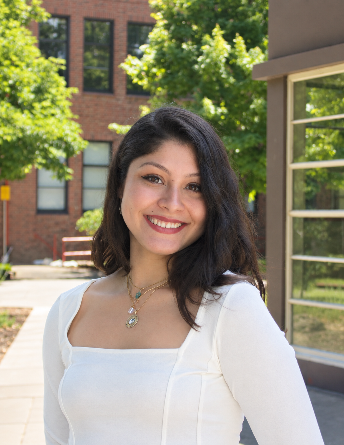 A smiling woman with dark hair and light skin standing outdoors on a sunny day, near a building with brick walls and green trees.