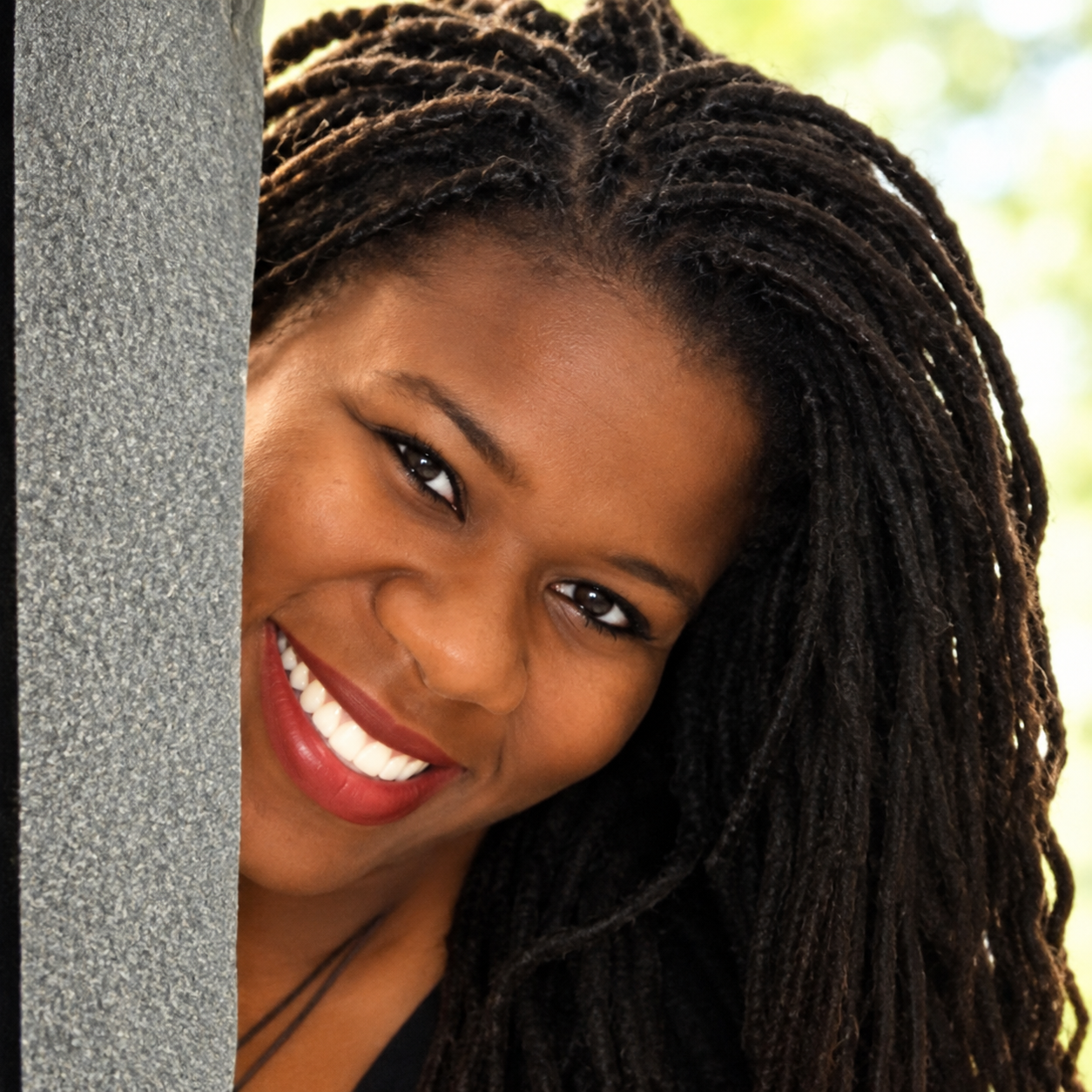 A smiling woman with dark curly hair peeking from behind a grey concrete pillar, outdoors with blurred green background.