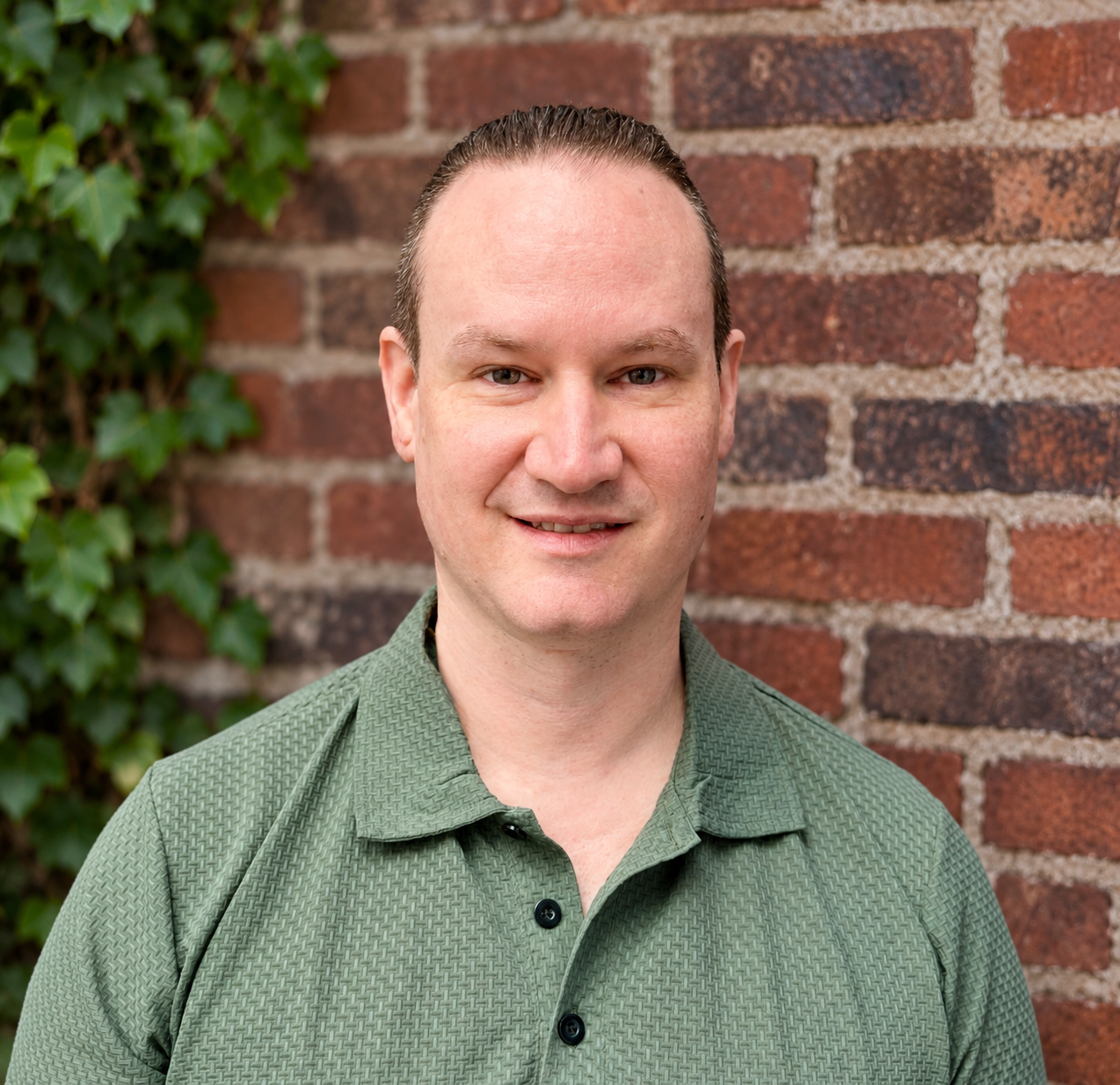 A man with short, dark hair, wearing a green polo shirt, standing in front of a brick wall with green ivy on the side, smiling at the camera.