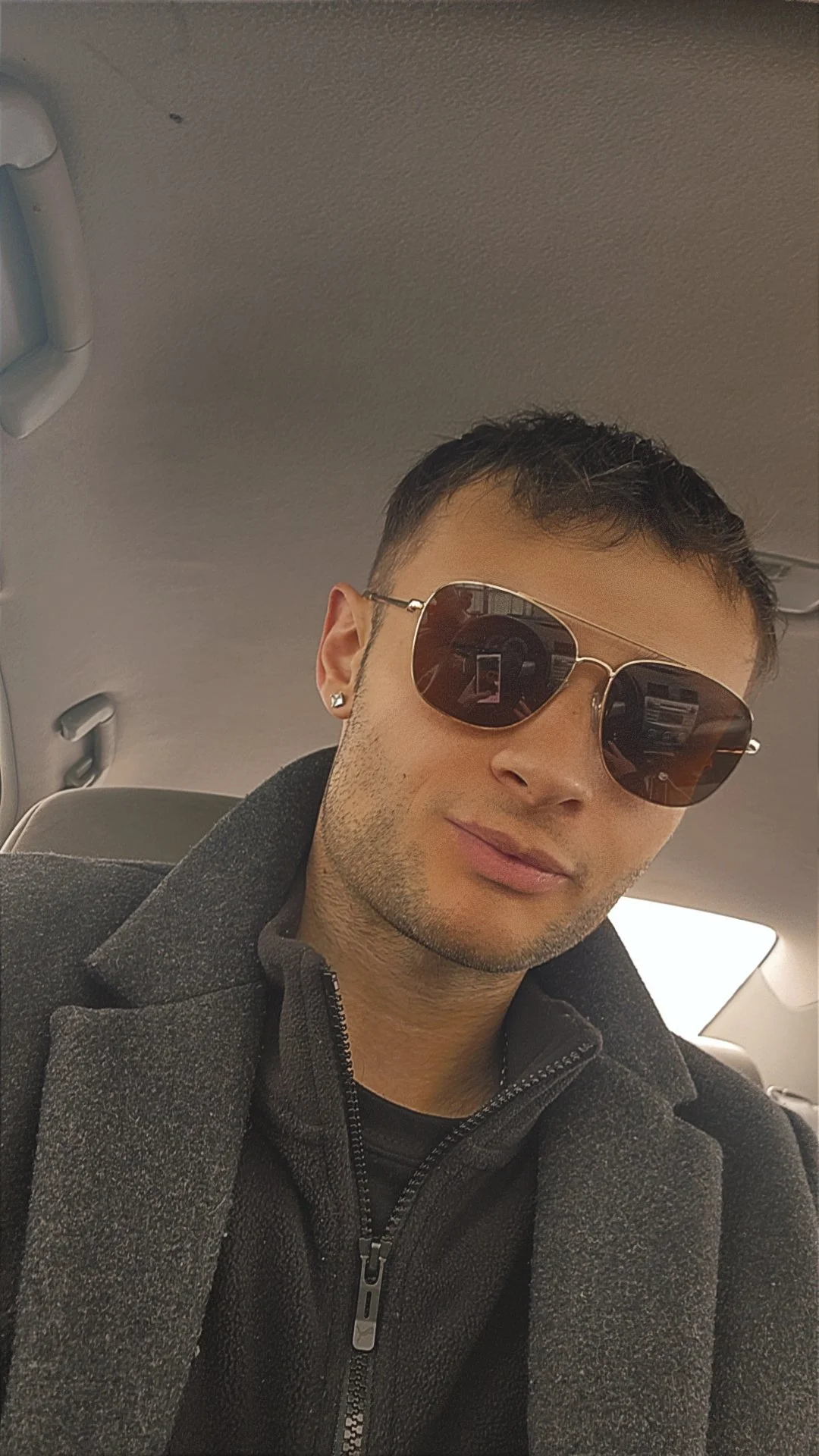 A young man with short dark hair, wearing aviator sunglasses and earrings, takes a selfie while sitting inside a vehicle. He is dressed in a dark zip-up jacket with a collar.